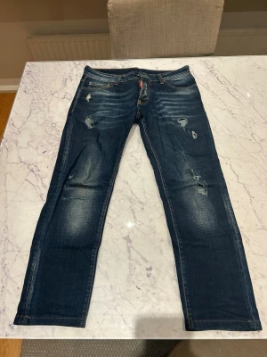 Dsquared2 Jeans - Snygga mörkblå jeans med raka ben och slitningar på flera ställen framtill. Jeansen har klassisk femficksdesign och ljusare partier vid lår och knän för en cool, använd look. Perfekta för en avslappnad streetstil.
