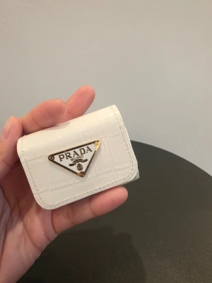 Vit AirPods-skal PD - Säljer en liten vit AirPods-skal från PD i läder. Väskan har ett trekantigt metallogo framtill och en silverfärgad kedja med karbinhake, perfekt som accessoar eller nyckelring.