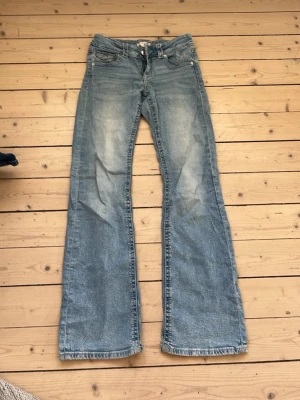 Ljusblå bootcut jeans med fickdetaljer - Säljer ett par ljusblå bootcut jeans med slitningar och snygga fickdetaljer bak med lock och knapp. Jeansen har klassisk femficksmodell och är tillverkade i denim med lätt tvättad look. Perfekta för en avslappnad och trendig stil.