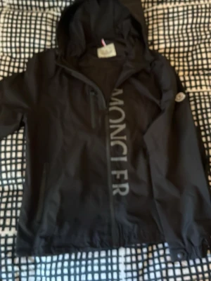 Svart vindjacka från Moncler - Snygg svart vindjacka från Moncler med huva och dragkedja framtill. Stora Moncler-bokstäver längs dragkedjan och logotyp på ärmen. Jackan har två sidofickor och justerbar nederkant. Perfekt för blåsiga dagar.