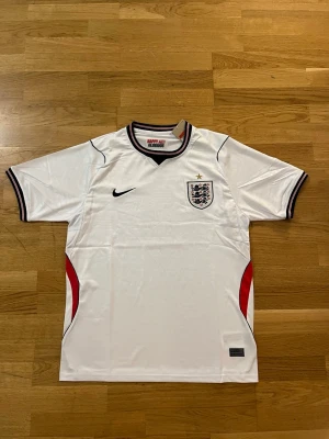  England fotbollströja vm - Säljer en vit England fotbollströja från Nike med korta ärmar. Tröjan har svarta och röda detaljer, broderad England-logga med stjärna på bröstet och texten 'HAPPY AND GLORIOUS' i nacken. Perfekt för fotbollsfans!     Fraktar samma eller dagen efter du köpt!!! Endast en kvar i lager!!!