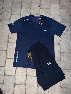 Marinblått träningsset från Under Armour - Säljer ett marinblått träningsset från Under Armour med t-shirt och shorts. Setet har logga på både tröja och shorts, samt färgglada detaljer och tryck. Materialet är lätt och andas, perfekt för träning. T-shirten har korta ärmar och rund hals.