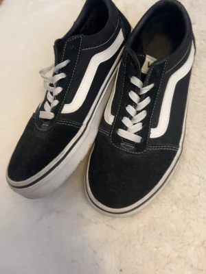 Svarta Vans Old Skool sneakers - Klassiska Vans Old Skool sneakers i svart mocka och canvas med vit sula och den ikoniska vita sidan. Skorna har vita snören och kontrastsömmar som ger en clean och tidlös look. Perfekta för dig som gillar streetwear och skate-stil.