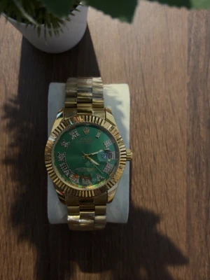 Klocka  - Stilren guldfärgad klocka med metallarmband och grön urtavla. Urtavlan har markerade siffror med stenar och datumvisning. Perfekt accessoar för dig som vill sticka ut med en lyxig look.