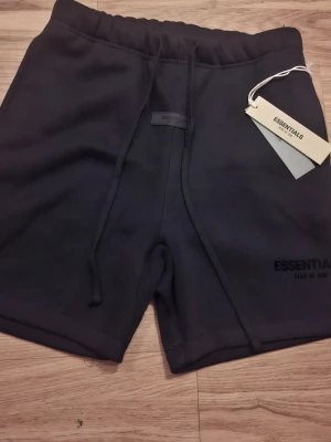 Svarta shorts från Essentials - Svarta shorts från Essentials med snörning i midjan och diskret logga på ena benet. Mjukt material och enkel design, perfekta för en avslappnad stil. Elastisk midja för skön passform.