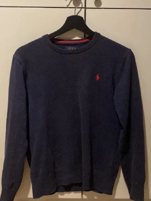 Mörkblå stickad tröja från Polo Ralph Lauren - Klassisk mörkblå stickad tröja från Polo Ralph Lauren med rund halsringning och den ikoniska röda logotypen broderad på bröstet. Tröjan har långa ärmar och ribbade muddar vid ärmslut och nederkant. Perfekt för en stilren och avslappnad look.