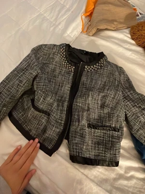 Grå kavaj med nitar vid kragen - Snygg gråmelerad kavaj med svart kantband och coola silverfärgade nitar runt kragen. Rak passform och två fickor framtill med svarta detaljer. Materialet är vävt tyg som ger en trendig look. Perfekt för dig som vill sticka ut lite extra. Kommer med ett helt set (med kjol)