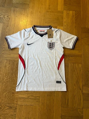England Nike vit matchtröja - Säljer en vit England matchtröja från Nike med svarta och röda detaljer. Tröjan har korta ärmar, V-ringad krage med svarta och röda ränder samt England-emblemet på bröstet. Perfekt för fotbollsfans som vill visa sitt stöd.