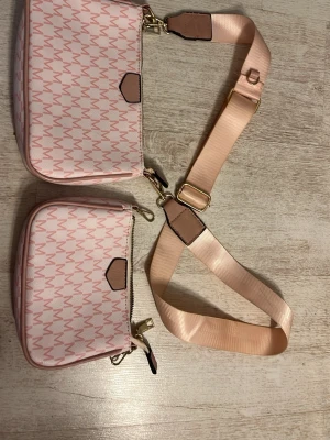Rosa axelväska med mönster - Säljer en snygg rosa axelväska med matchande mindre väska. Väskorna har ett M-mönster i ljusrosa och vita toner, justerbar axelrem i satinliknande material och guldfärgade detaljer. Perfekt för dig som vill ha både stil och funktion i ett.
