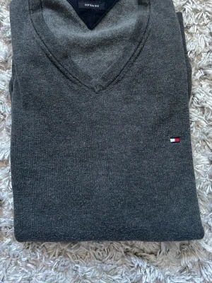 Grå stickad tröja Tommy Hilfiger - Mörkgrå stickad tröja från Tommy Hilfiger i mjuk bomull/silkesblandning. Tröjan har V-ringning och den klassiska lilla Hilfiger-loggan broderad på bröstet. Perfekt för dig som gillar stilrena och tidlösa plagg. Väldigt bra skick.