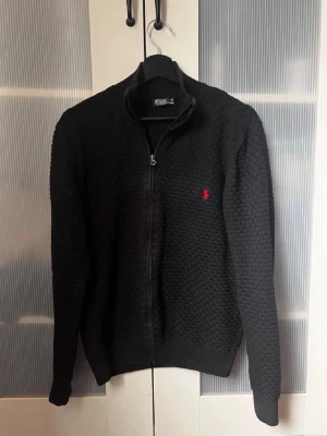 Svart ziptröja från Polo Ralph Lauren - Svart stickad ziptröja från Polo Ralph Lauren med diskret röd logga på bröstet. Tröjan har en strukturerad yta, hög krage och hel dragkedja framtill. Perfekt för lager-på-lager och har ribbade muddar vid ärmslut och nederkant.