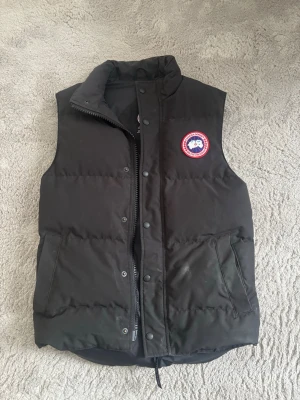 Svart dunväst från Canada Goose - Snygg svart dunväst från Canada Goose med klassisk logga på bröstet. Västen har hög krage, dragkedja och tryckknappar framtill samt två stora fickor. Perfekt för lager-på-lager och håller dig varm under kyliga dagar.