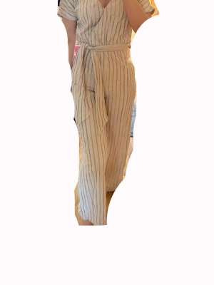 Beige randig jumpsuit med knytband - Säljer en beige jumpsuit med tunna vertikala ränder i vitt och brunt. Modellen har korta ärmar, omlott-ringning och ett matchande knytband i midjan. Tillverkad i ett lätt och luftigt tyg, perfekt för varma dagar. Vida ben ger en avslappnad och trendig look.