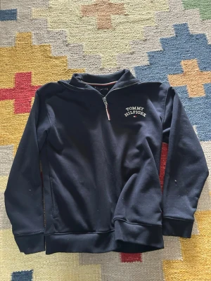 Svart halfzip hoodie Tommy Hilfiger - Snygg svart hoodie från Tommy Hilfiger med halv dragkedja och broderad logga på bröstet. Tröjan har ribbade muddar vid ärmslut och nederkant samt en klassisk, stilren look. Perfekt för dig som gillar streetwear och vill ha något bekvämt och trendigt.