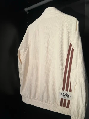Vit track jacket från VAG RAN - Vit track jacket i strukturerat tyg med dragkedja framtill och två fickor. Bruna bokstäver 'VAG RAN' på bröstet och tre bruna ränder längs ena ärmen. Liten patch med texten 'Vuitton' på ryggen. Sportig och avslappnad stil, perfekt för streetwear.