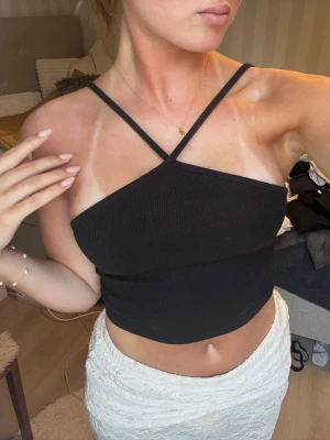 Svart ribbad croppad linnetopp - Snygg svart croppad linnetopp med ribbad struktur och tunna axelband som korsas framtill. Perfekt till sommarens outfits och lätt att matcha med både kjol och byxor. Materialet känns mjukt och följsamt mot huden.