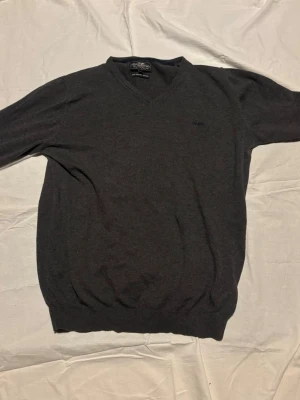 Mörkgrå stickad tröja Hampton Republic - Mörkgrå stickad tröja från Hampton Rebulic med V-ringning och diskret broderad logga på bröstet. Tröjan har långa ärmar och ribbade muddar vid ärmslut och nederkant. Perfekt för lager på lager och enkel att matcha.