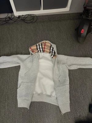 Grå Burberry hoodie med dragkedja - Snygg grå hoodie från Burberry med klassiskt rutigt foder i huvan och dragkedja framtill. Hoodien har två fickor och ribbade muddar. Perfekt för en chill och stilren look. Materialet är mjukt och bekvämt, och huvan har dragsko.