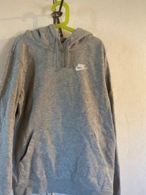 Grå hoodie från Nike med huva - Säljer en klassisk grå hoodie från Nike med vit logga på bröstet. Hoodien har huva med dragsko och en stor magficka framtill. Tillverkad i mjukt bomullsmaterial som är skönt att ha på sig. Perfekt för chill dagar eller när du vill ha en enkel och snygg look.