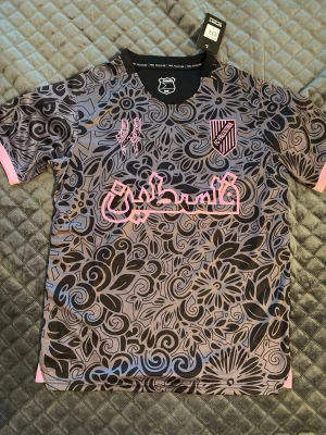 PC Palestina fotbollströja mönstrad - Unik fotbollströja från PC Palestina i storlek S. Tröjan har ett grått och svart blommigt mönster med rosa detaljer, tryck och emblem. Rund halsringning och korta ärmar. Tillverkad i lätt och ventilerande material, perfekt för träning eller match.