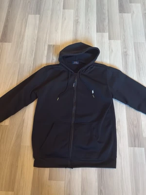 Svart hoodie från Polo Ralph Lauren - Svart hoodie med dragkedja från Polo Ralph Lauren. Klassisk design med huva och dragsko, två fickor framtill och den ikoniska lilla vita loggan broderad på bröstet. Perfekt för en chill och stilren look.
