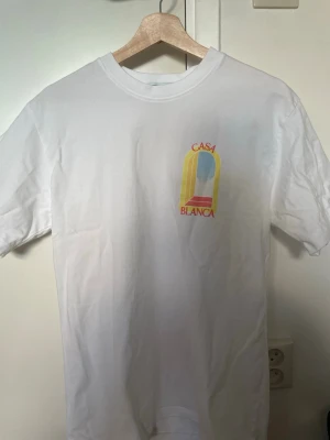 Vit Casa Blanca t-shirt med tryck - Vit t-shirt från Casa Blanca med färgglatt tryck på bröstet i gult, blått och rött. Klassisk rund hals och korta ärmar. Perfekt för dig som gillar stilrena plagg med en twist. Materialet känns mjukt och skönt mot huden.
