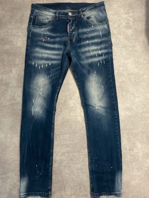 Dsquared2 blå skinny jeans med färgstänk - Snygga blå skinny jeans från Dsquared2 med coola slitningar och färgstänk i vitt och rött. Jeansen har klassisk femficksdesign, knappgylf och en röd Dsquared2-tag vid gylfen. Perfekt för dig som gillar en trendig och edgy stil.