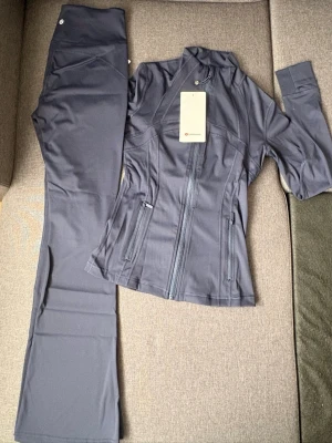 Mörkgrått träningsset från Lululemon - Snyggt mörkgrått träningsset navy från Lululemon med figursydd jacka och matchande byxor. Jackan har dragkedja framtill, ståkrage och två fickor med dragkedja. Byxorna är raka och stretchiga, perfekta för gym eller yoga. Priset kan diskuteras 🌸💖🌷 labels are not available in the item.