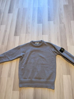 Beige stickad tröja från Stone Island - Snygg beige stickad tröja från Stone Island med klassisk rund halsringning och ribbade muddar. Tröjan har den ikoniska svarta logotypen med kompass på vänster ärm. Perfekt för dig som gillar stilrena och trendiga plagg.