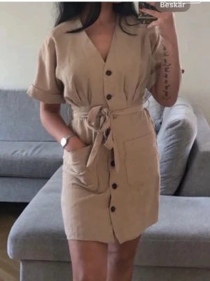 Zara Beige kort klänning med knytskärp - Stilren beige kortklänning med v-ringning, knappar framtill och knytskärp i midjan. Klänningen har korta ärmar med uppvik och stora fickor framtill. Perfekt för en avslappnad och trendig look.