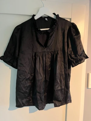 Svart blus med volangkrage - En svart blus med lätt glansigt tyg, volangdetalj vid kragen och korta puffärmar med resår vid ärmslut. Lös passform och lätt rynk under bysten ger en avslappnad look. Perfekt att styla med jeans eller kjol.
