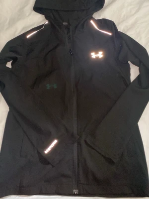 Svart träningsjacka från Under Armour - Snygg svart träningsjacka från Under Armour med huva och hel dragkedja. Reflekterande detaljer på axlar och logga framtill samt texten UNDER ARMOUR på ärmen. Perfekt för löpning eller utomhusträning. Tillverkad i ett lätt och funktionellt material.