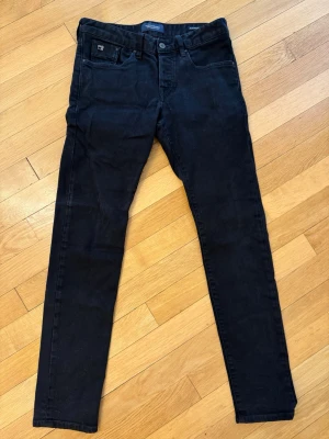 Svarta slim fit jeans från Scotch & Soda - Snygga svarta slim fit jeans från Scotch & soda med klassisk femficksdesign och knappgylf. Jeansen har raka ben och är tillverkade i en mjuk bomullsblandning med lite stretch för extra komfort. Perfekta till en avslappnad streetwear-look. Använda men fortfarande i bra skick!
