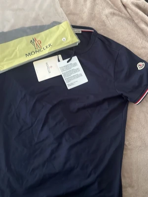 Marinblå Moncler t-shirt - Snygg marinblå t-shirt från Moncler med korta ärmar och Moncler-logga på ärmen. Klassisk rund halsringning och diskreta rödvita detaljer vid ärmslut. Tillverkad i mjuk bomull för en skön känsla. Perfekt för dig som gillar stilrena plagg med exklusiv touch.