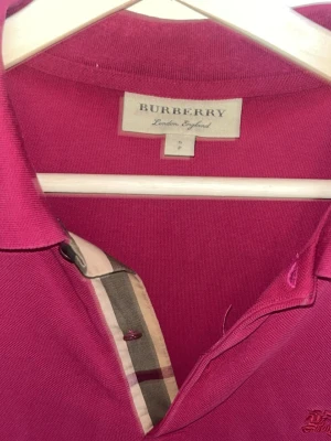 Rosa pikétopp från Burberry - Säljer en rosa pikétopp från Burberry med klassisk krage och knappslå med det ikoniska rutiga Burberry-mönstret. Toppen har broderad logga på bröstet och är tillverkad i mjuk bomull. Perfekt för en stilren och trendig look.