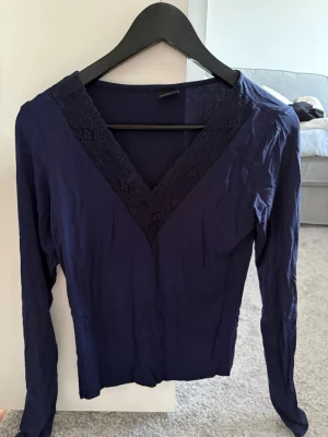 Mörkblå topp med spetsdetalj - Mörkblå blus med djup V-ringning och dekorativ spets längs urringningen. Långärmad modell i mjukt tyg som ger en stilren och elegant look. Använd fåtal gånger
