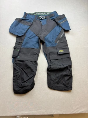 Arbetsbyxa Snickers Workwear blå/svart - Säljer ett par slitstarka arbetsbyxor från Snickers Workwear i blått och svart. Byxorna har flera praktiska fickor, förstärkta knän och smarta detaljer för verktyg. Materialet är tåligt och designen är anpassad för rörlighet och funktion.