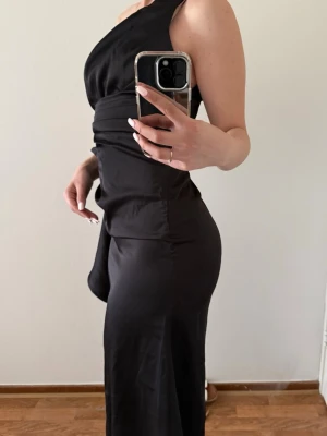 Svart asymmetrisk långklänning - Säljer en svart långklänning med asymmetrisk, one shoulder-design och draperad midja. Klänningen har en elegant siluett och är tillverkad i ett mjukt, följsamt tyg som ger en snygg passform. Perfekt för dig som vill ha en stilren och trendig look.