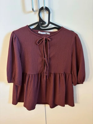 Plommonlila blus från 157 med knyt - Trendig plommonlila blus från lager 157 i storlek XS. Blusen har trekvartsärm, rund halsringning och snygg knytning framtill. Tillverkad i ett mjukt, ribbat tyg som ger en avslappnad men ändå chic look. Perfekt att styla med jeans eller kjol.