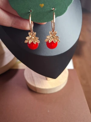 Guldörhängen med röda pärlor - Snygga örhängen med guldfärgade blommor och klarröda pärlor som hänger ned. Örhängena har en elegant och trendig design med fina detaljer i metall och färgstarka pärlor. Perfekta för att ge din look en färgklick och lite extra bling.