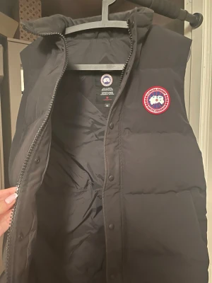 Svart dunväst från Canada Goose - Snygg svart dunväst från Canada Goose med klassisk logotyp på bröstet. Västen har hög krage, dragkedja och tryckknappar framtill. Tillverkad i slitstarkt material och fylld med dun för extra värme. Perfekt för lager-på-lager och streetwear-stil.
