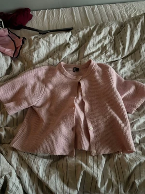 Kort rosa kofta från H&M - Söt kort kofta i ljusrosa från H&M med ribbad struktur och korta ärmar. Koftan har rund halsringning och vita knappar framtill. Perfekt att slänga över en topp för en chill look.