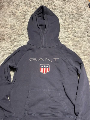 Mörkblå hoodie från GANT - Snygg mörkblå hoodie från GANT med stor huva och känguruficka. Framtill finns GANT-loggan och ett broderat emblem med stjärnor och ränder i rött, vitt och blått. Perfekt för en avslappnad och sportig stil. Tillverkad i mjukt bomullsmaterial.