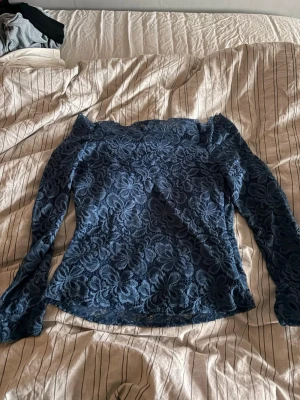 Blå spetsblus med blommönster - Säljer en snygg blå långärmad blus i spets med blommigt mönster. Toppen har en figurnära passform och är perfekt för dig som gillar detaljer. Offshoulder-designen ger en trendig touch och materialet känns lätt och luftigt.