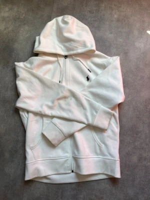 Vit hoodie från Polo Ralph Lauren - Snygg vit hoodie med dragkedja från Polo Ralph Lauren. Klassisk design med huva, fickor framtill och den ikoniska lilla loggan broderad på bröstet. Perfekt för en clean och avslappnad stil. Mjuk bomullskänsla och ribbade muddar.