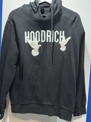 Svart hoodie från Hoodrich med duvor - Svart hoodie från Hoodrich med stor vit logga och två broderade duvor på bröstet. Klassisk huva med dragsko och ribbade muddar. Perfekt för dig som gillar streetwear och vill sticka ut med snygga detaljer.
