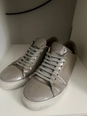 Gråa sneakers från Axel Arigato - Snygga gråa sneakers från Axel Arigato med blank finish och vita sulor. Skorna har klassisk låg design, matchande grå skosnören och diskret logga på sidan. Tillverkade i läder för en clean och stilren look. Unika och sällsynta pris kan diskuteras, påse medföljer. Lite smuts som kan tas bort av en enkel tvätt.