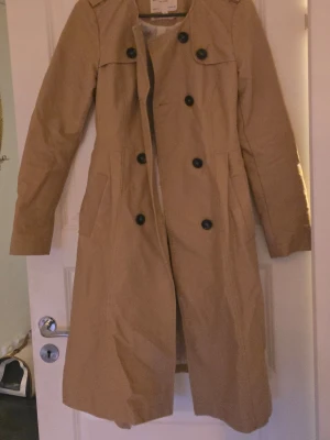 Beige trenchcoat från Holly & Whyte - Stilren beige trenchcoat från Holly & Whyte by Lindex. Klassisk dubbelknäppt design med svarta knappar, axelklaffar och fickor på sidorna. Lång modell i ett lätt tyg, perfekt för vår och höst.