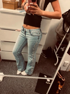Ljusblå bootcut jeans med brodyr - Säljer ett par ljusblå bootcut jeans med snygg vit blommig brodyr längs utsidan av benen. Jeansen har klassisk femficksmodell och låg midja. Perfekta för dig som vill ha en unik och trendig look.💋 längd 88 midjemått 36