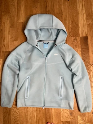 Ljusblå hoodie från Nike NOCTA - Snygg ljusblå hoodie från Nike NOCTA med dragkedja framtill och två fickor med dragkedja. Hoodien har en vit Nike-logga på bröstet och en skön, sportig känsla. Perfekt för dig som gillar streetwear och vill ha något stilrent och bekvämt.